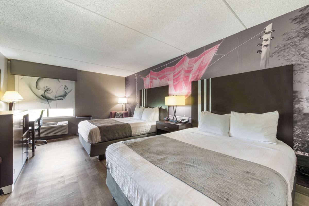 Une chambre d’hôtel avec deux lits, un bureau avec une chaise, une lampe et un grand tableau abstrait accroché au mur.