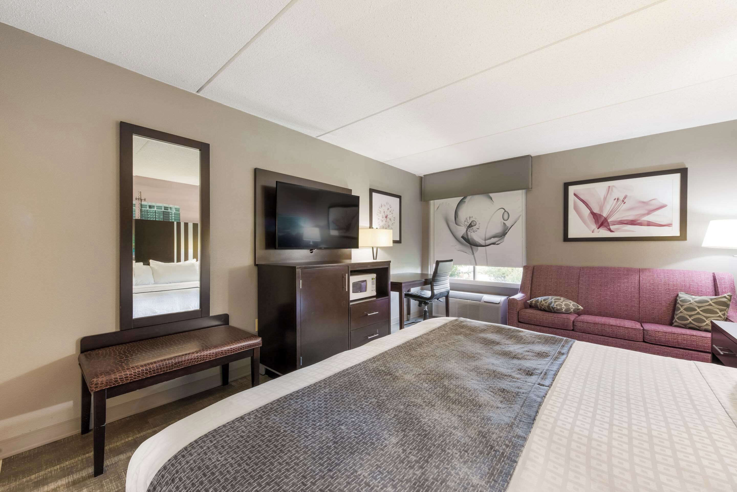 Una habitación de hotel con una cama grande, un escritorio con computadora, televisor, banca, sofá y obras de arte en las paredes.