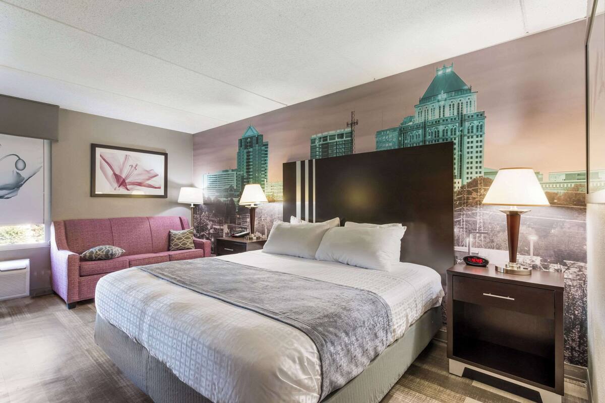 Une chambre d’hôtel avec un grand lit, un canapé rose, deux tables de chevet avec des lampes et une fresque représentant un paysage urbain sur le mur.