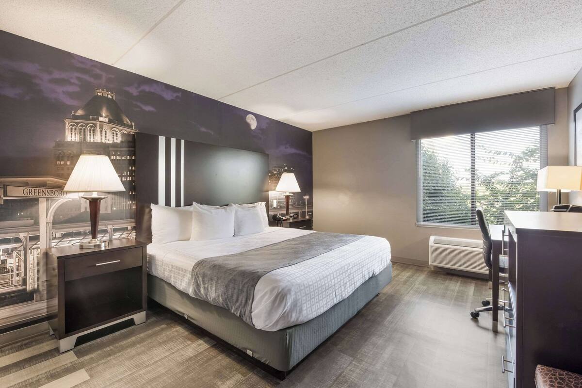 Une chambre d’hôtel avec un grand lit, deux tables de chevet avec des lampes, un bureau avec une chaise et une fenêtre avec des stores.