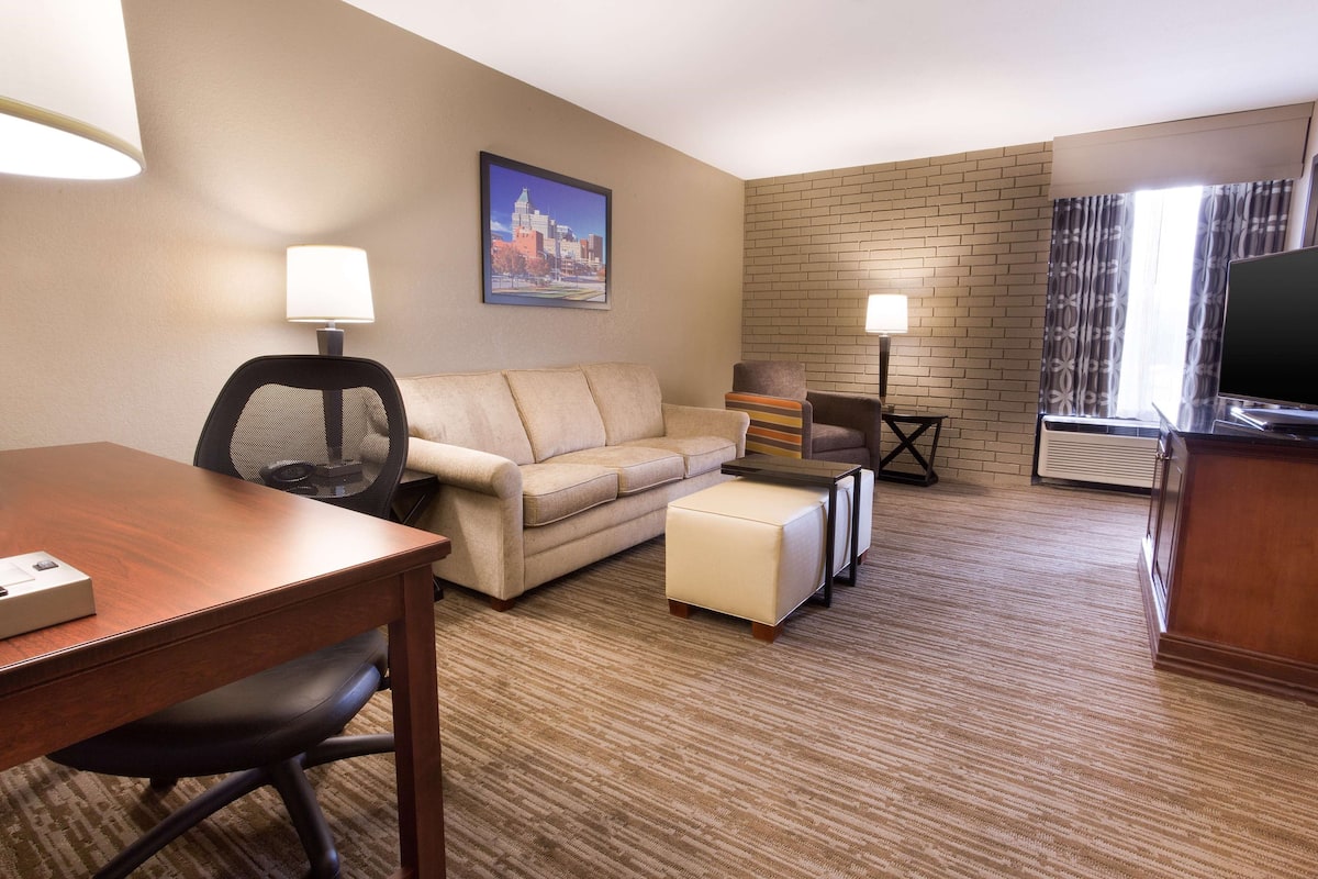 A hotel room with a wooden desk, beige sofa, a small ottoman, a TV, and a cityscape painting on the wall.