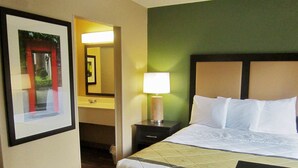 Habitación de hotel con cama, mesita de noche, lámpara, un cuadro enmarcado y un baño visible a través de una puerta abierta.