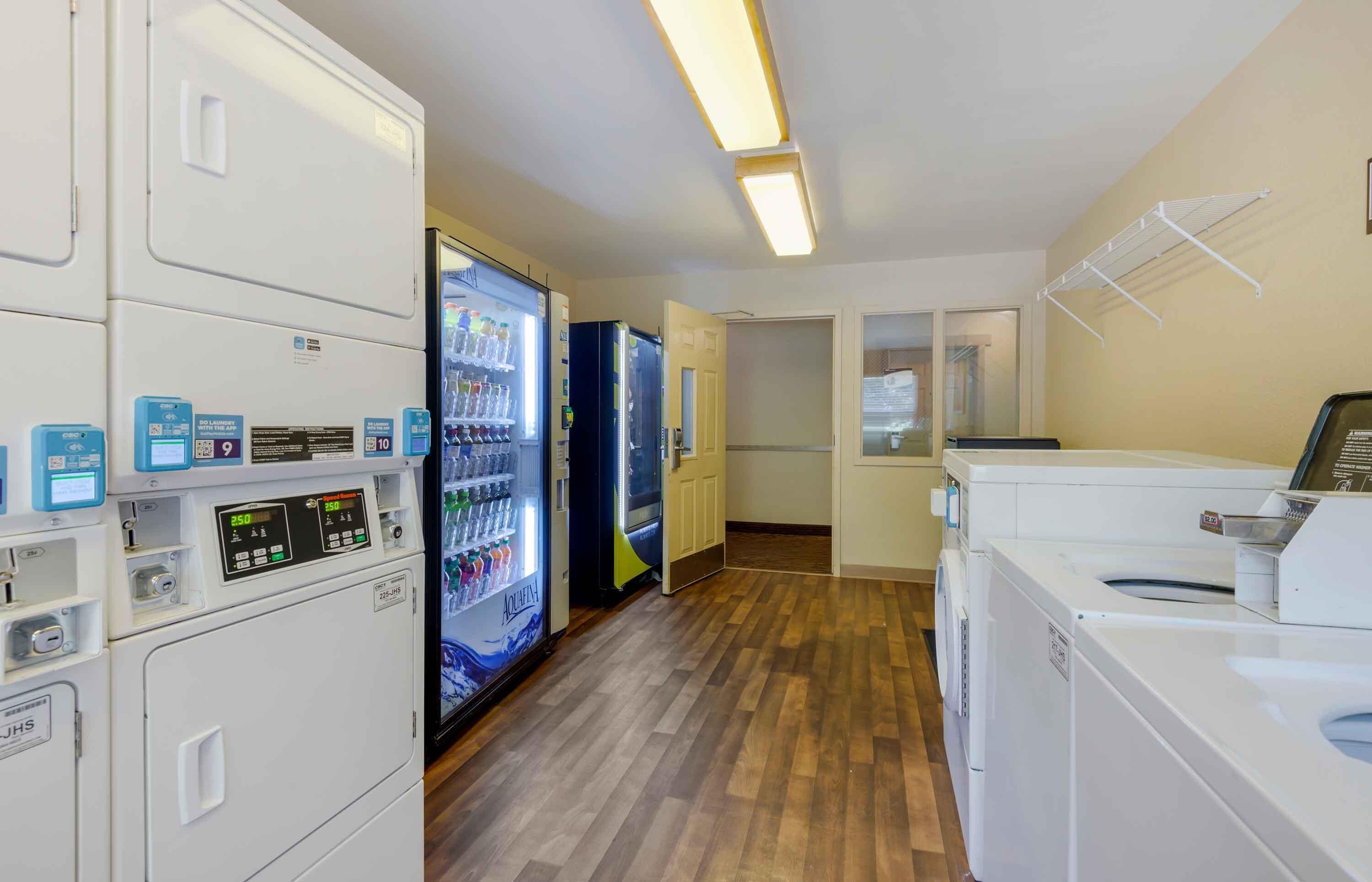 A laundry facility with multiple washing machines and dryers, a refrigerator with beverages, and a door leading to another room.