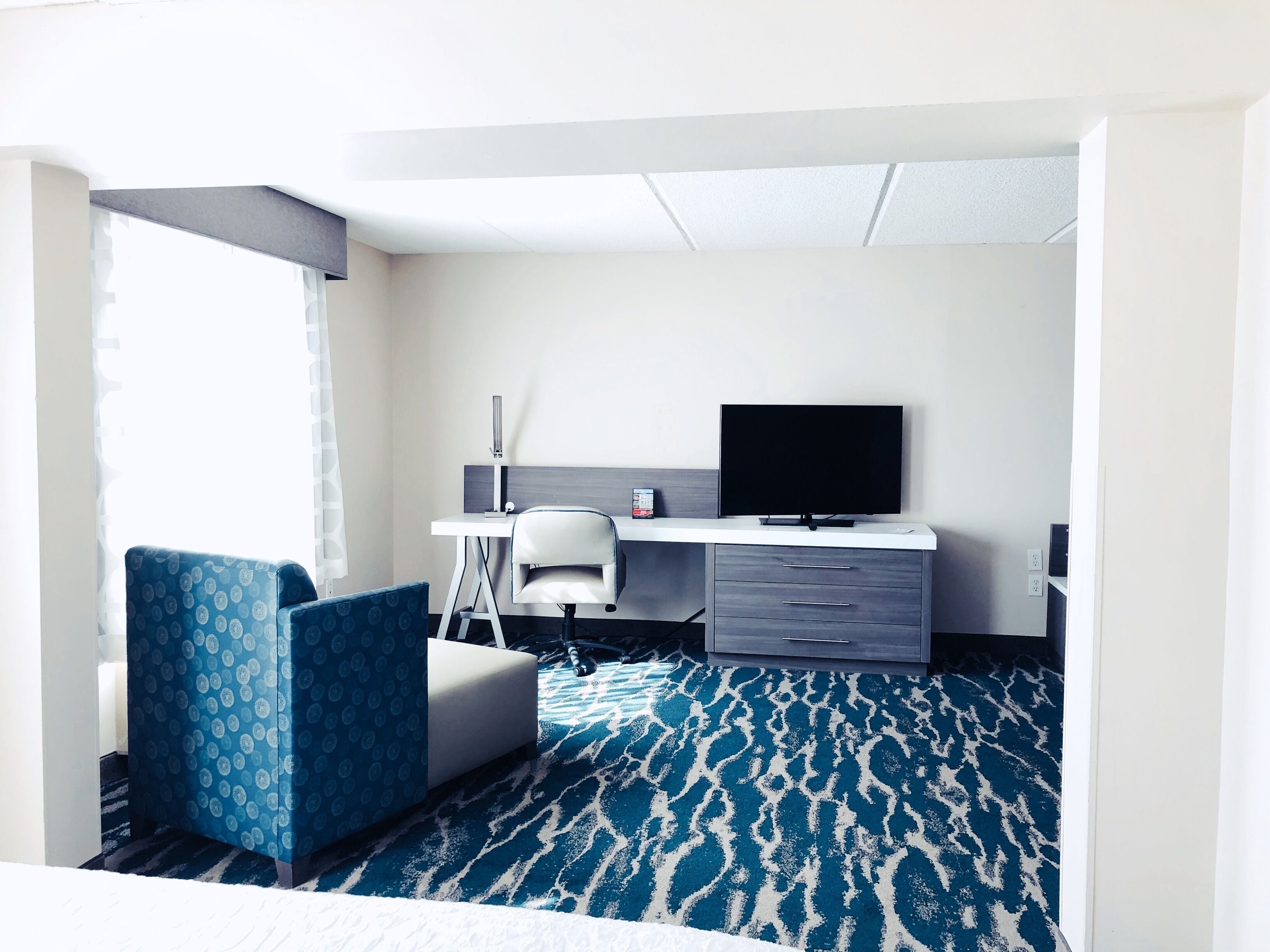 Habitación de hotel con cama, escritorio con computadora, silla, televisor y alfombra estampada.