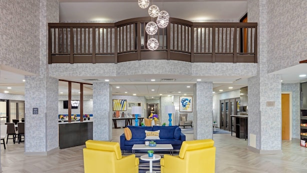 Un hall d’entrée spacieux, avec un canapé bleu, des fauteuils jaunes, une table basse centrale et une balustrade en bois.