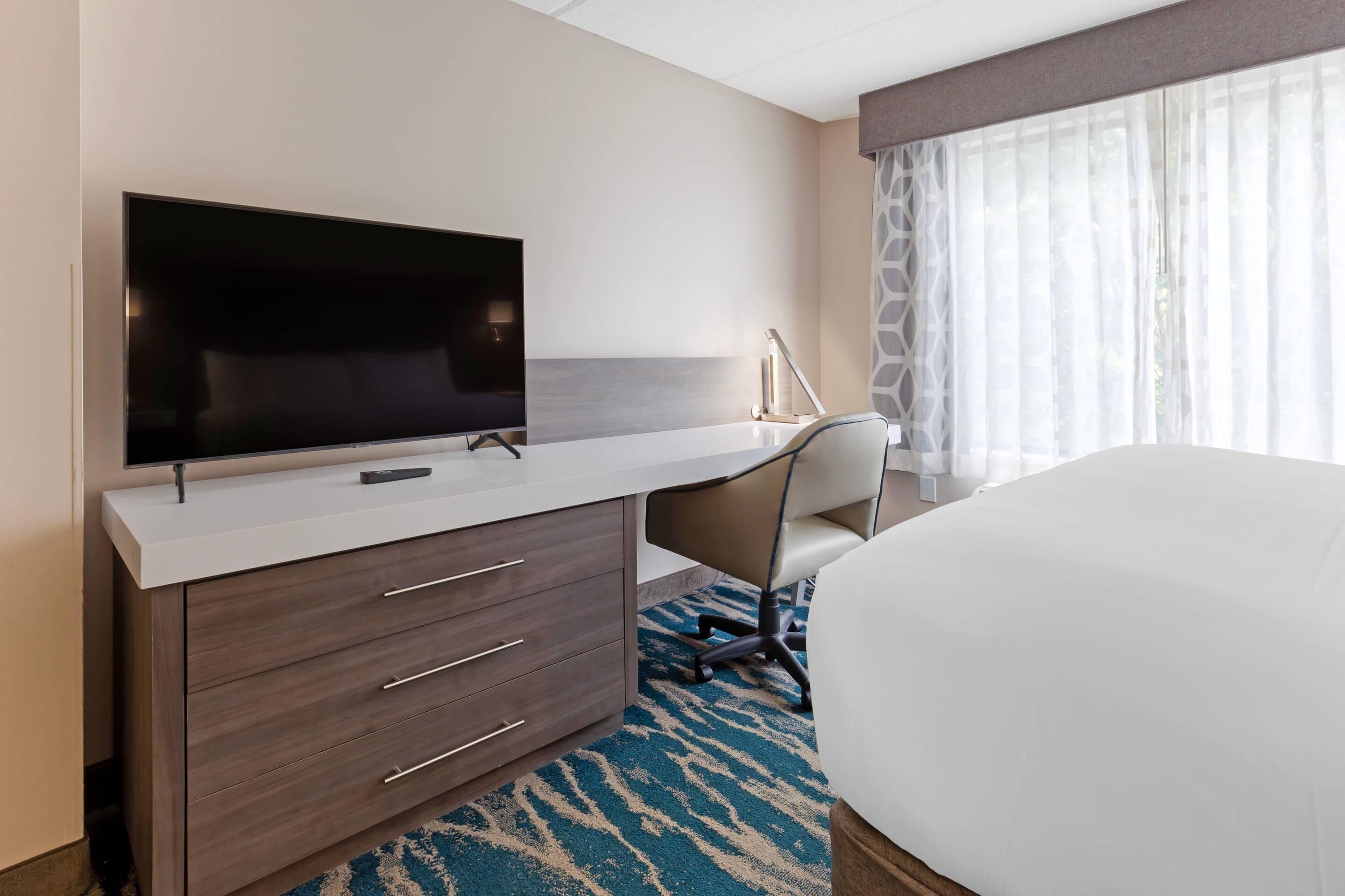 Quarto de hotel com cama, escrivaninha com televisão de tela plana, cadeira e janela grande com cortinas finas.