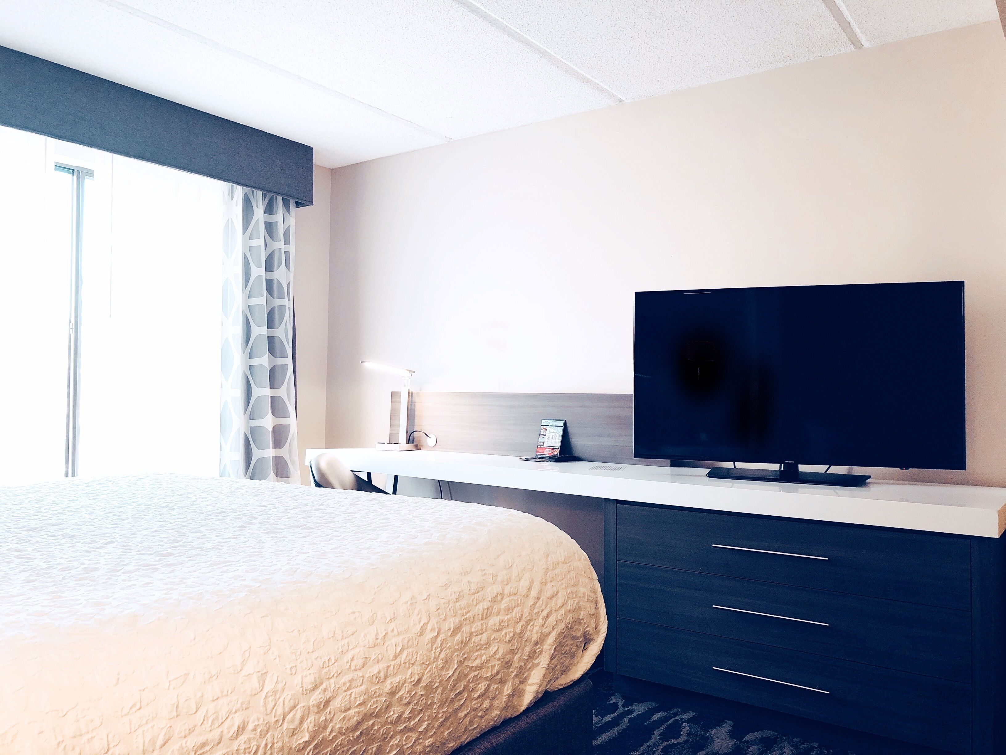 Habitación de hotel con cama, escritorio, televisor de pantalla plana, lámpara y ventana con cortinas.