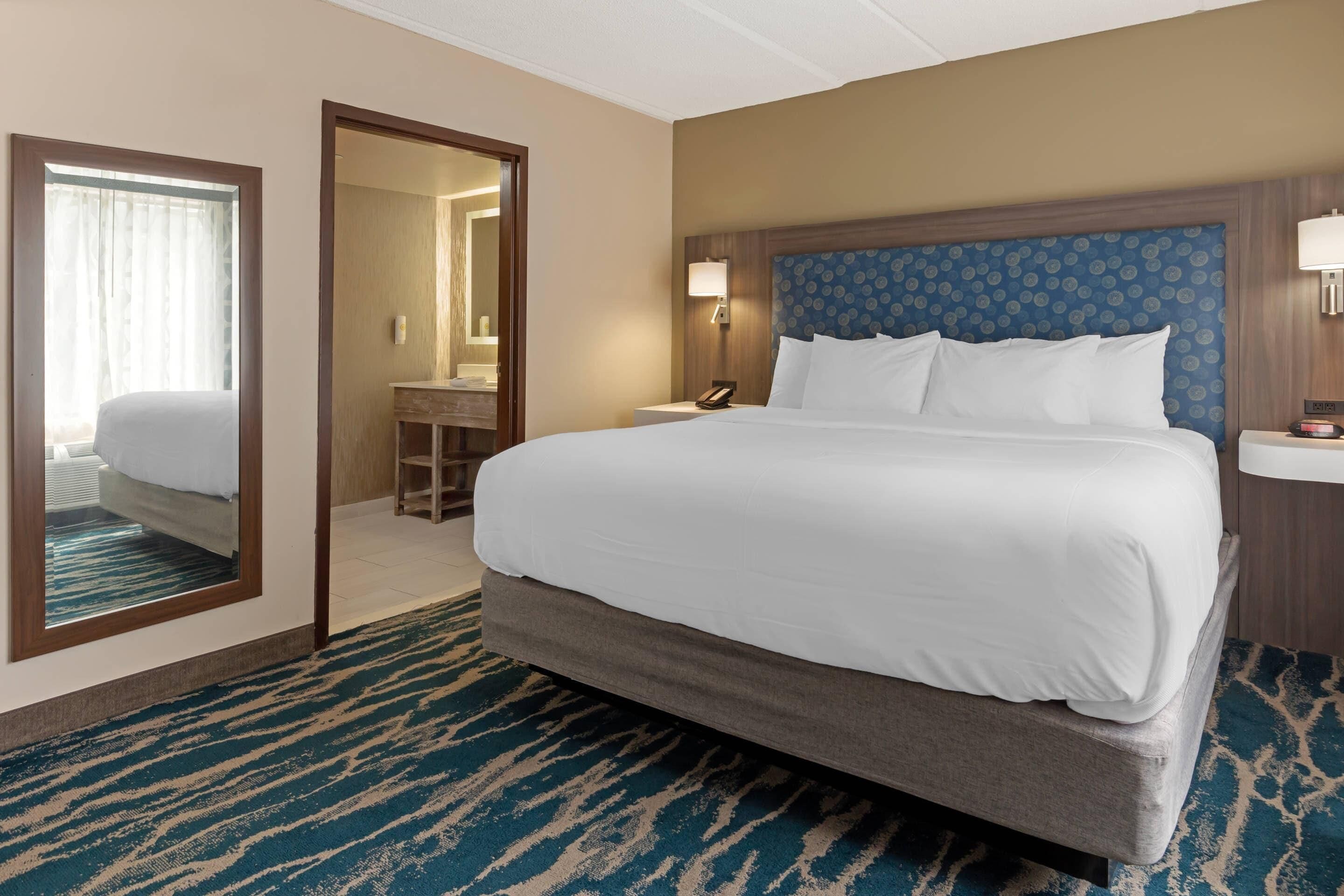 Een hotelkamer met een groot bed, een spiegel, een nachtkastje met een lamp en een badkamer zichtbaar door een geopende deur.