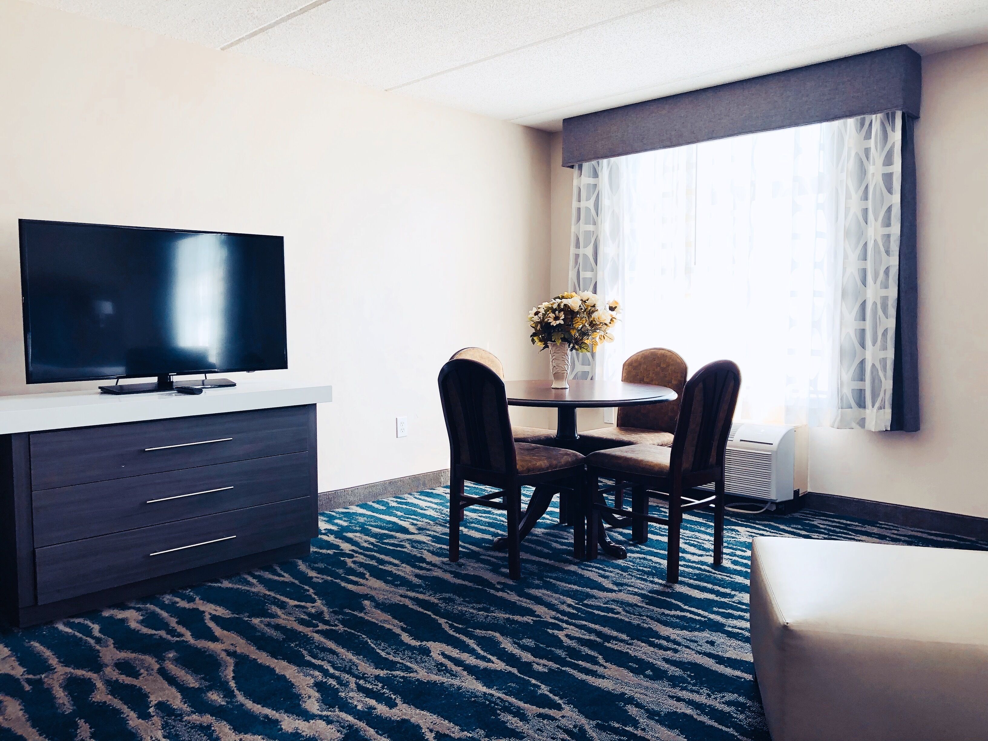 A hotel room with a flat-screen TV on a white console, a dining table with four chairs, a vase with flowers, and a large window with patterned curtains.