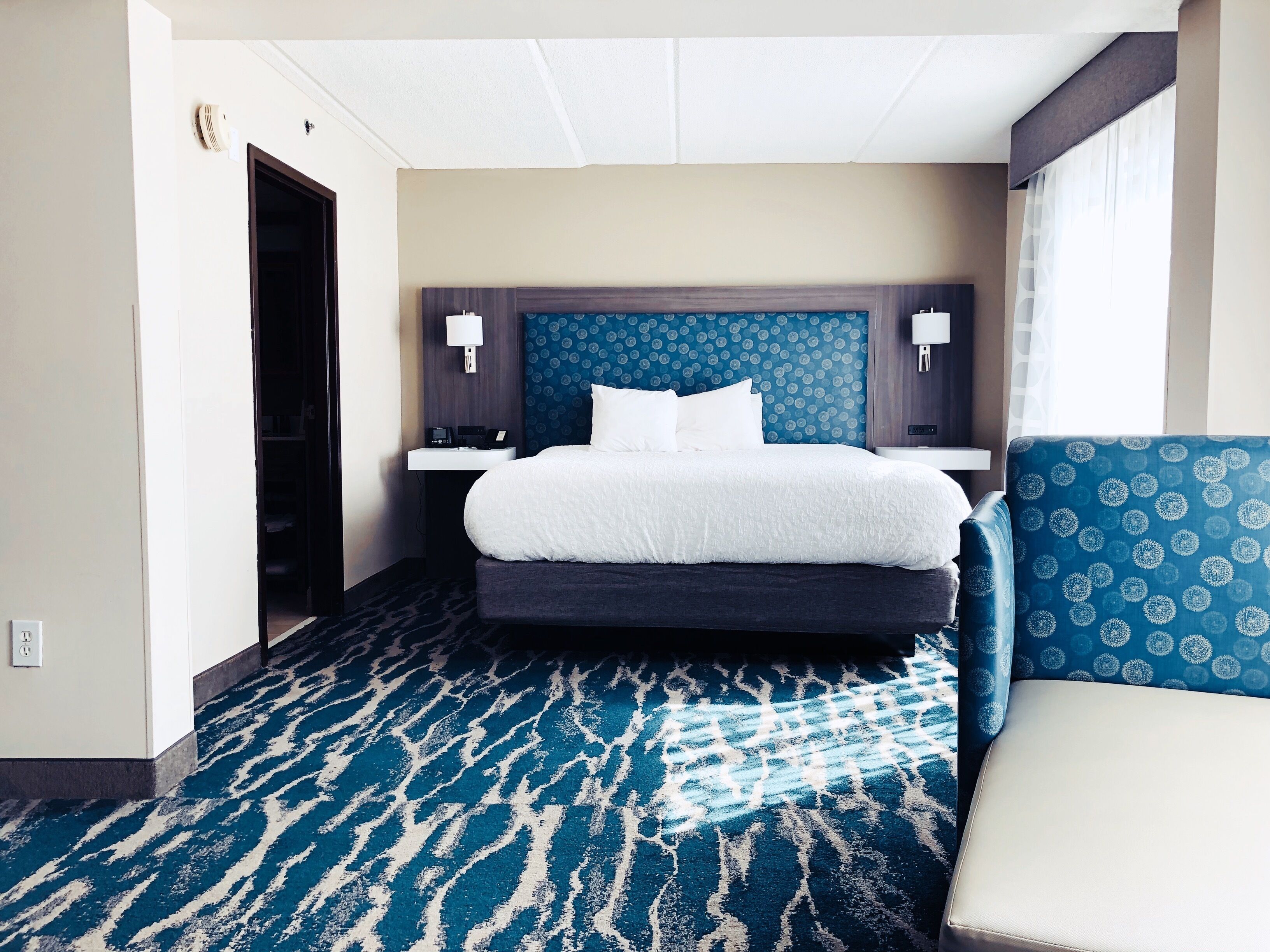 Habitación de hotel con una cama grande, una mesita de noche, una silla, alfombra estampada y un purificador de aire montado en la pared.