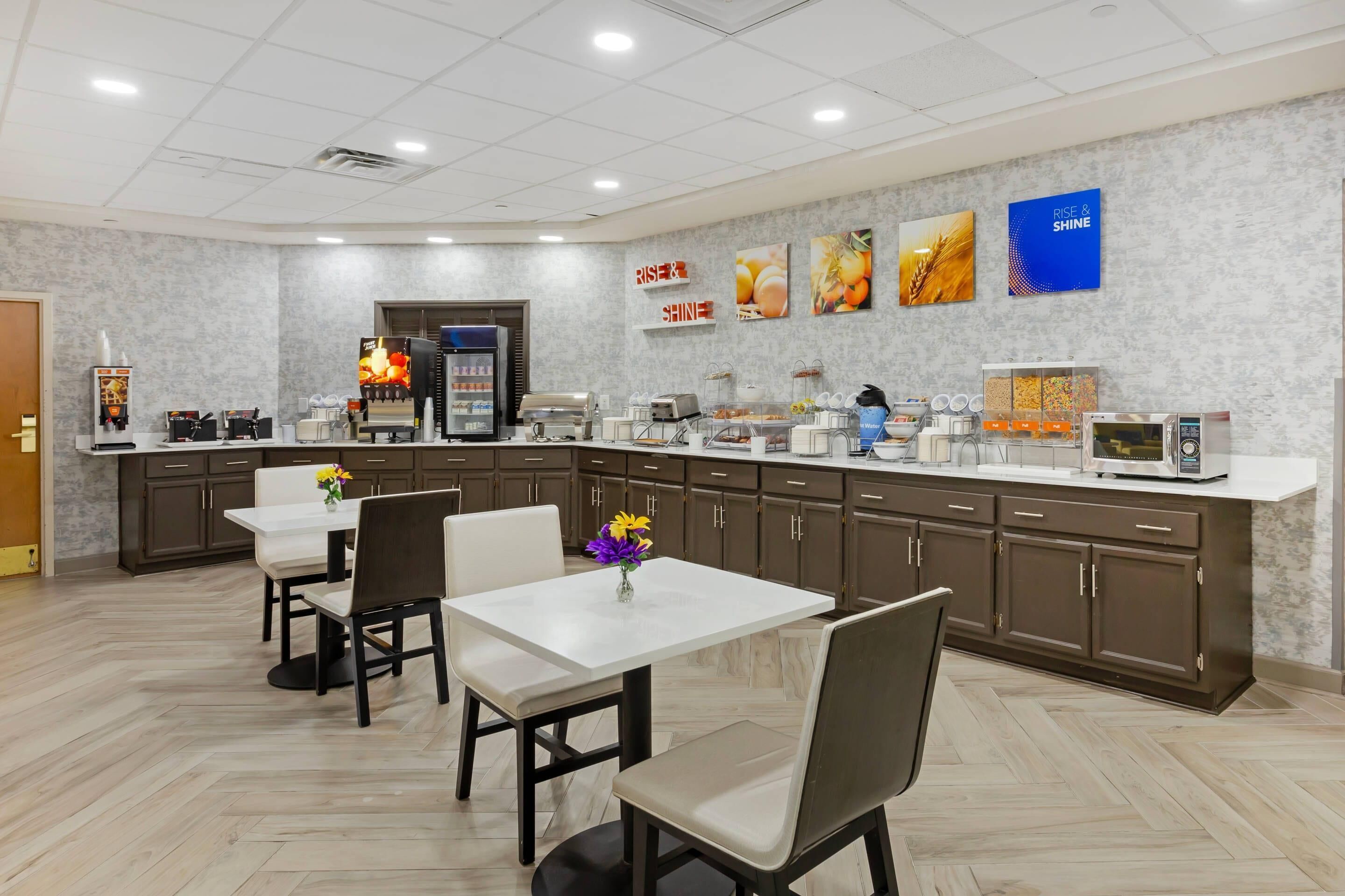 A modern dining area with a long counter, several tables, and chairs. There are food service stations and a refrigerator in the background.