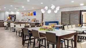 Un espace repas moderne avec plusieurs tables et chaises, une cuisine en arrière-plan et un luminaire décoratif.