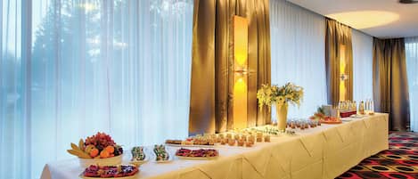 A long banquet table with various food items and a vase of flowers, set against a backdrop of large windows with sheer curtains.