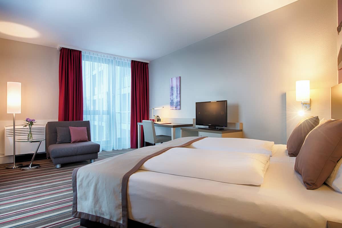 Una habitación de hotel con una cama grande, un escritorio con televisor, un sofá y una ventana con cortinas.