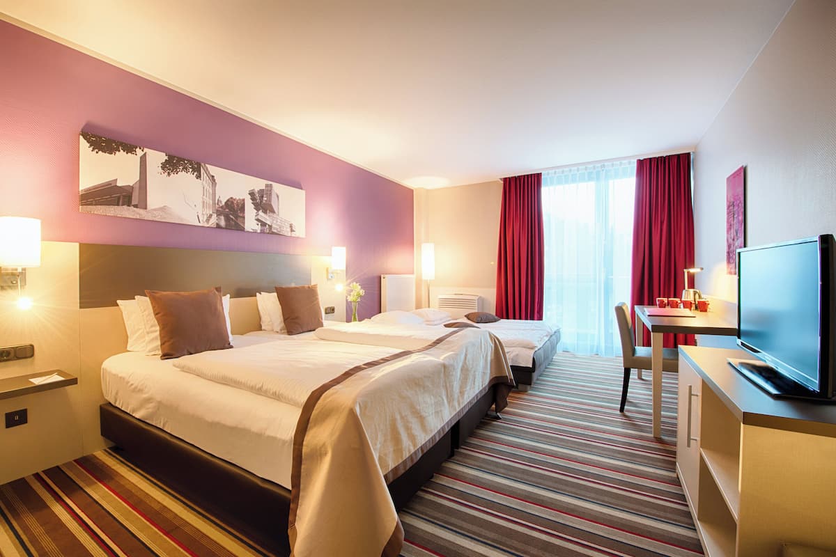 Habitación de hotel con dos camas, un escritorio con televisor y un ventanal con cortinas rojas.