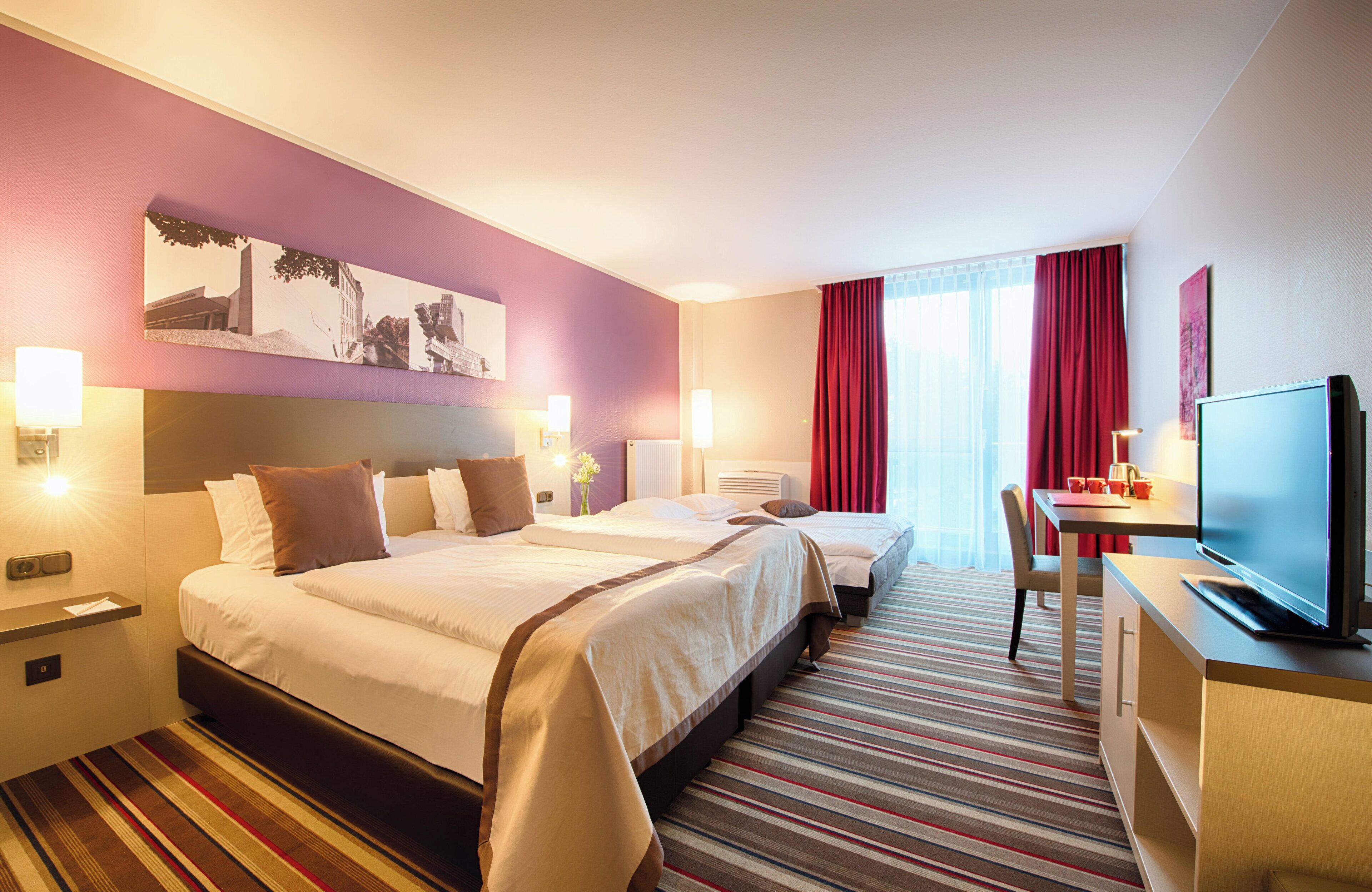 Habitación de hotel con dos camas, un escritorio con televisor y un ventanal con cortinas rojas.