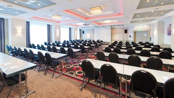 Un salón de conferencias con filas de mesas y sillas, una pantalla de proyector y piso alfombrado.