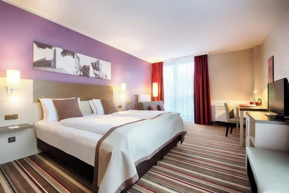 Habitación de hotel con una cama grande, mesitas de noche, un escritorio con una silla, un televisor y una ventana con cortinas.