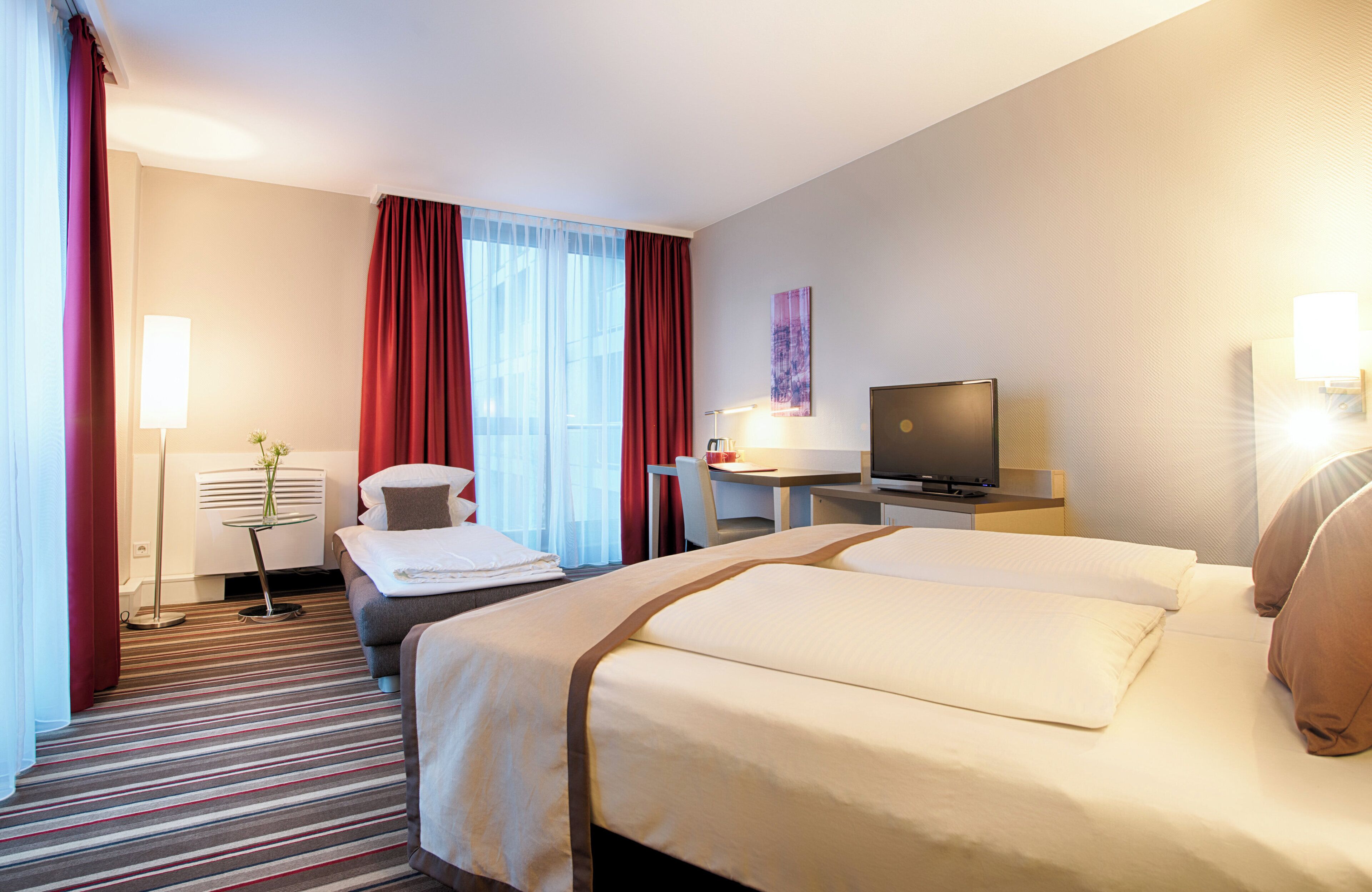 A hotel room with two beds, a desk, a chair, a TV, and a lamp.