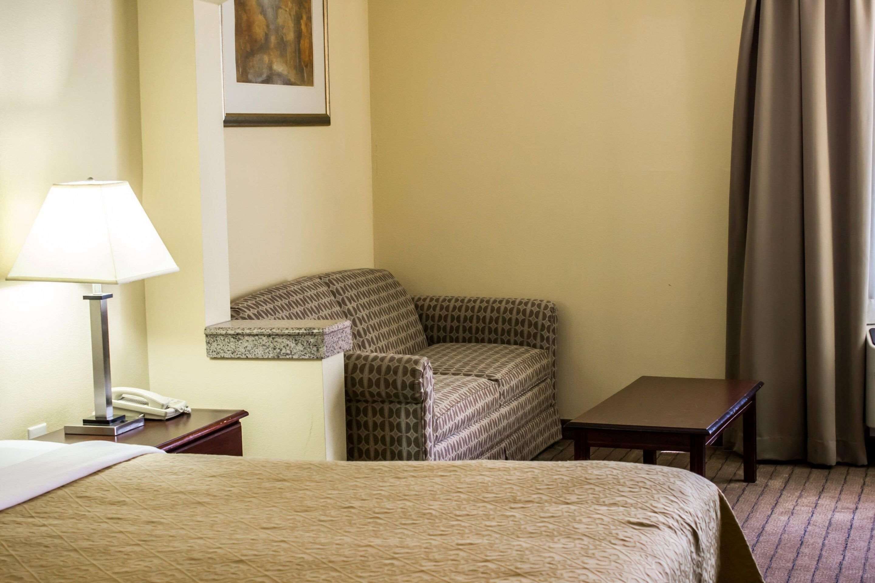 A hotel room with a bed, a chair, a small table, a lamp, and a phone on a nightstand.