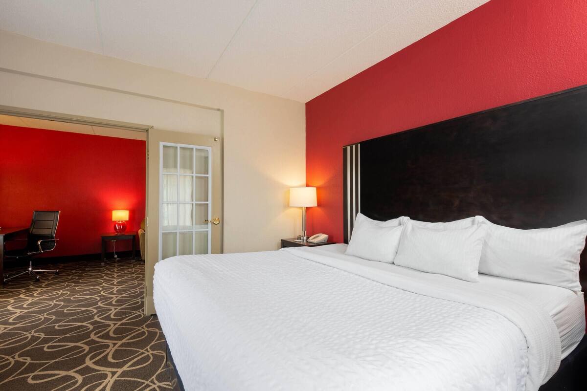 Een hotelkamer met een groot bed, een bureau met stoel, een lamp en een deur naar een andere kamer.
