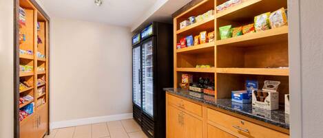 A kitchenette with wooden cabinets, a refrigerator, and shelves stocked with snacks and beverages.