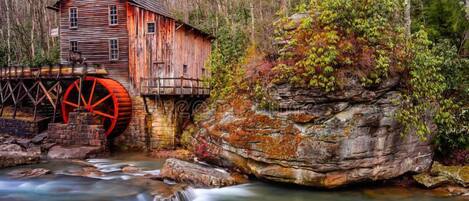 A rustic wooden mill with a large water wheel, set in a forested area by a flowing river.