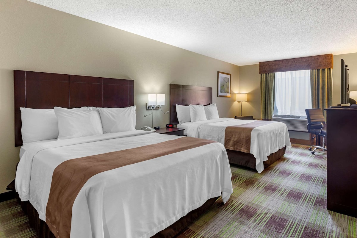 A hotel room with two beds, a desk, a chair, a lamp, and a window with curtains.