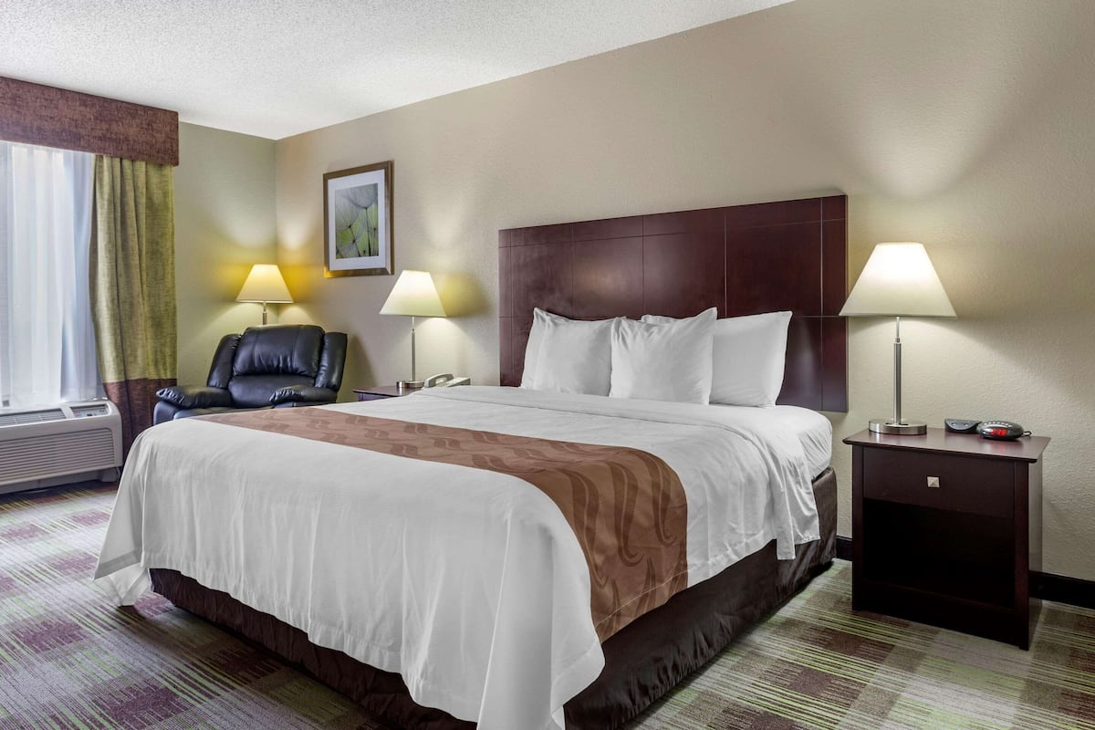 A hotel room with a large bed, two bedside lamps, a leather chair, and a nightstand with a clock.