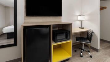 A modern hotel room with a flat-screen TV mounted on a wooden shelf, a black refrigerator, a yellow storage unit, a desk with a lamp, and a black office chair.