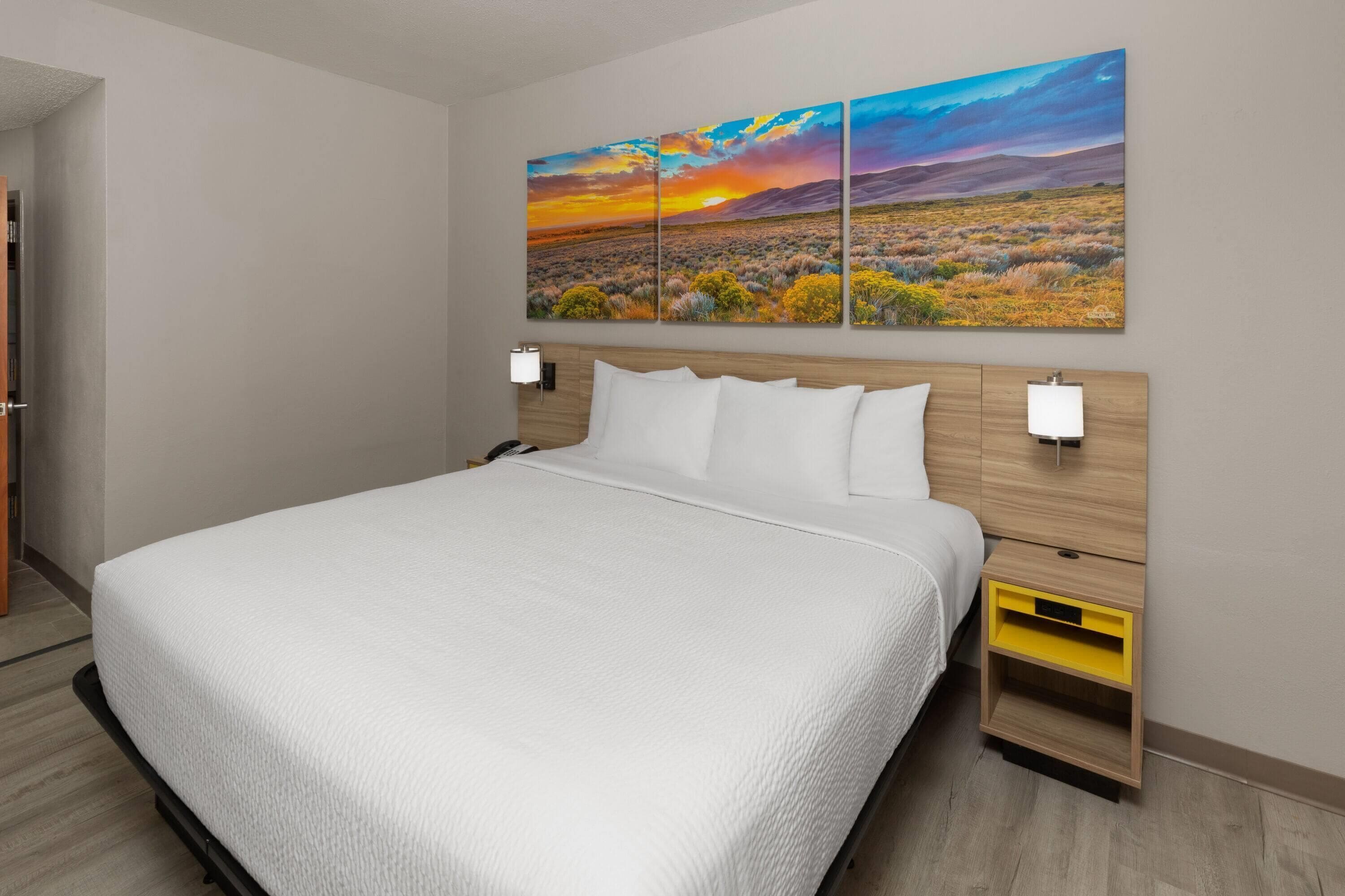 A hotel room with a large bed, a wooden headboard, a nightstand with a lamp, and a wall-mounted painting of a sunset landscape.