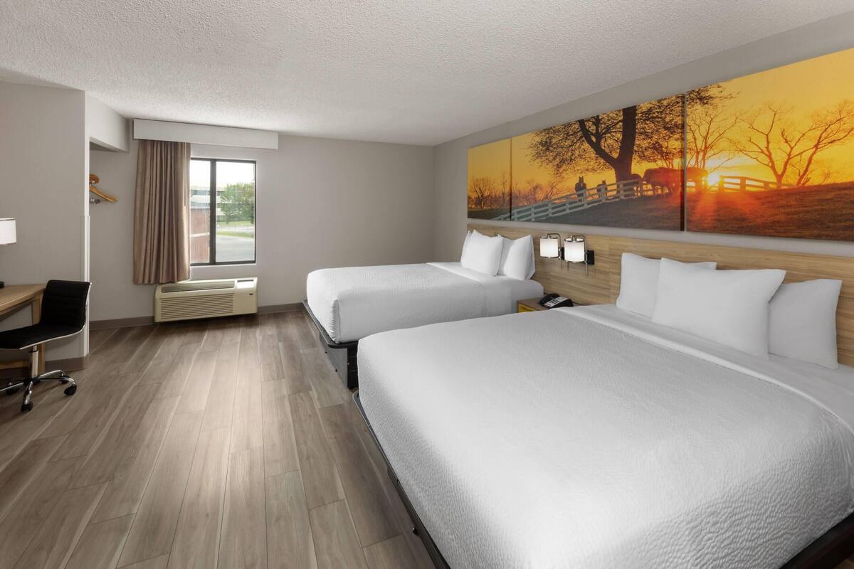 Une chambre d’hôtel avec deux lits, un bureau, une chaise, une fenêtre avec des rideaux et un grand tableau au mur.