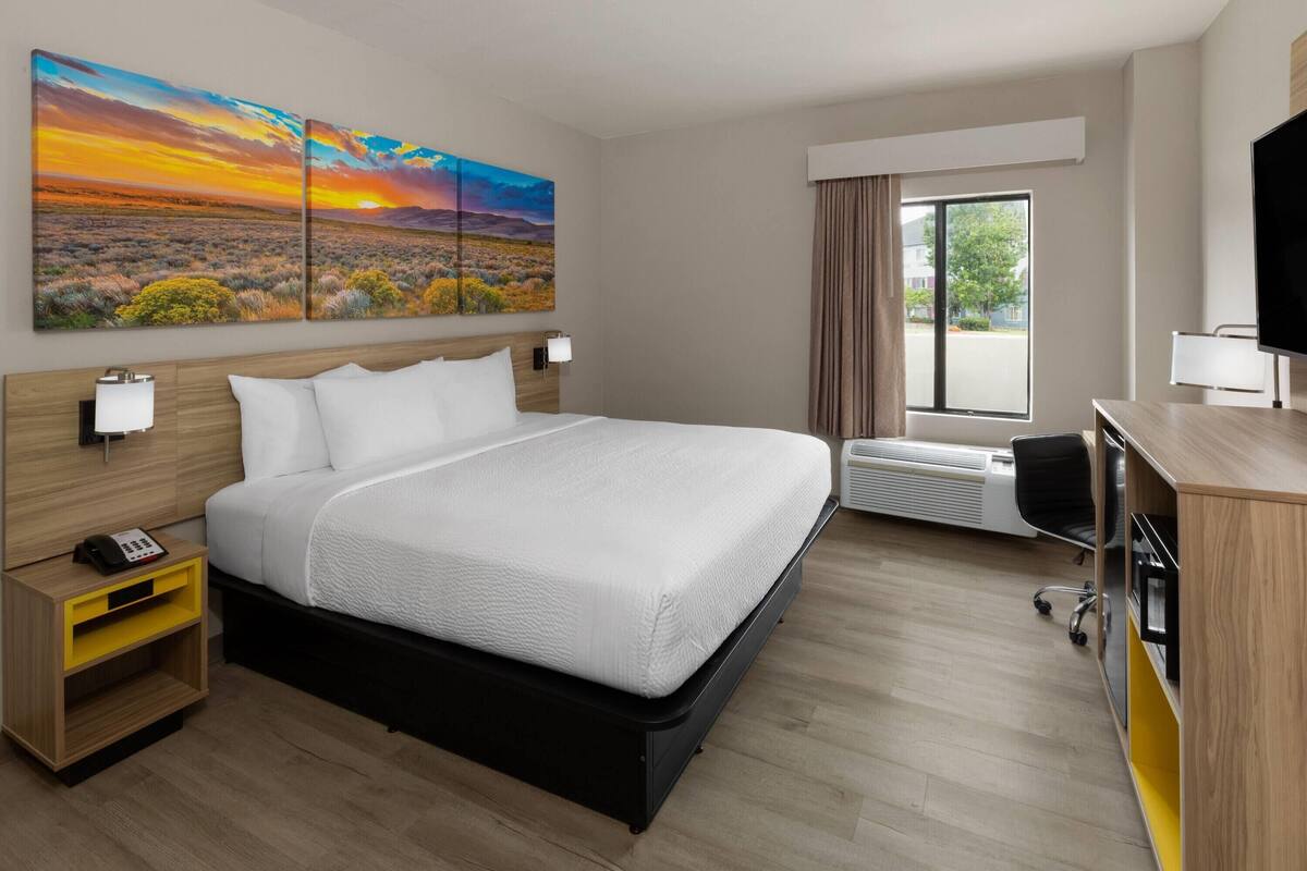Une chambre d’hôtel avec un grand lit, un bureau, une chaise, une télévision, une fenêtre avec des rideaux et deux tableaux au mur.