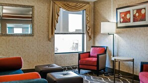 A hotel room with a large window, a mirror, a red armchair, a blue armchair, a small table, and a floor lamp.