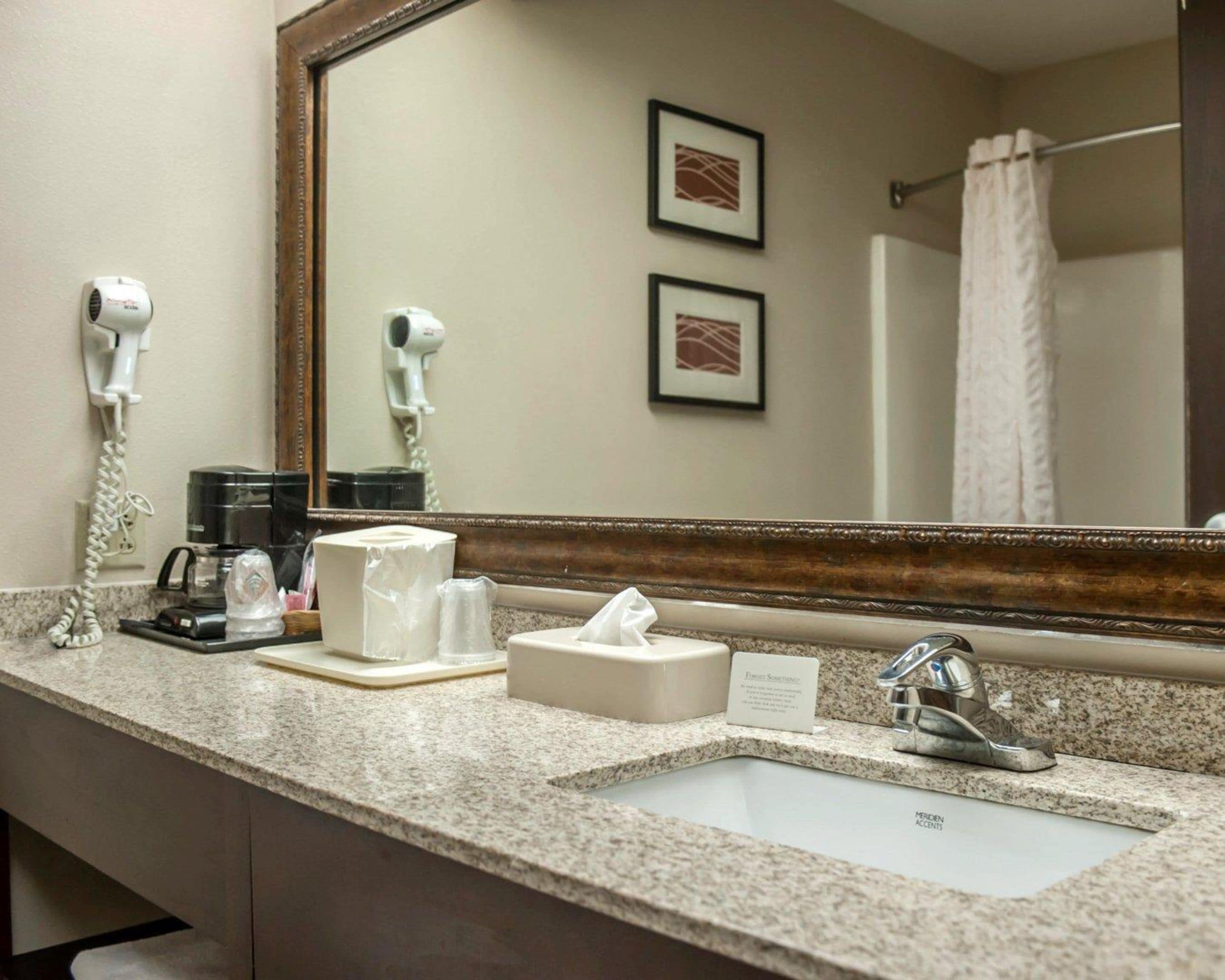 A hotel bathroom with a granite countertop, a sink, a mirror, a hairdryer, a coffee maker, and a towel rack.
