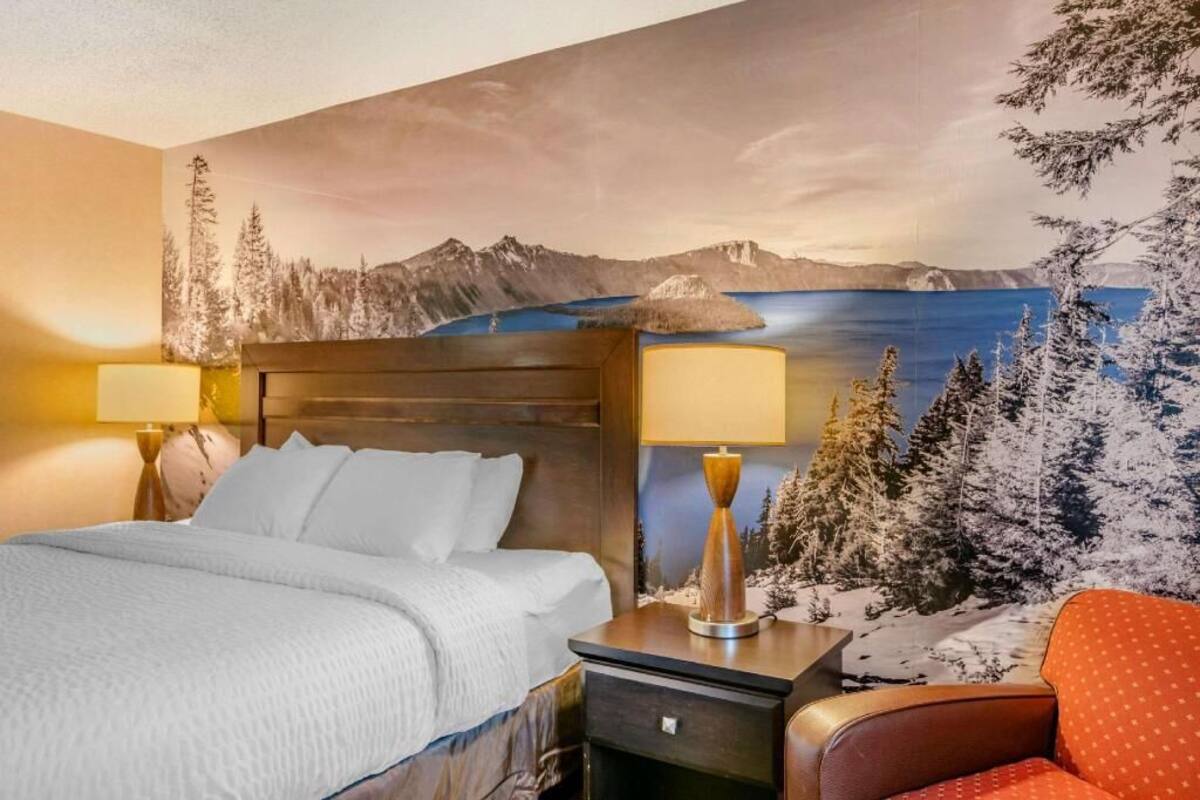 Quarto de hotel com cama, mesas de cabeceira, uma cadeira e um mural de paisagem montanhosa.