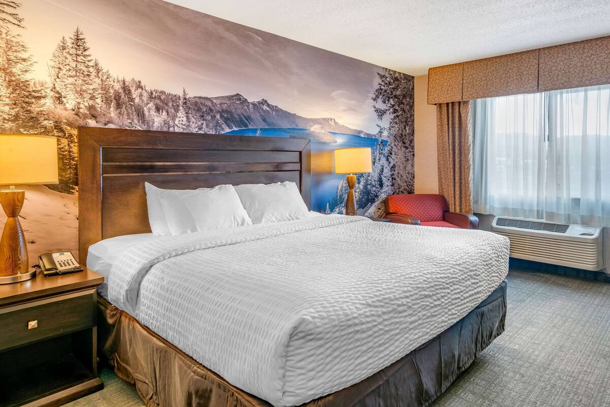 Uma cama grande com cabeceira de madeira, duas luminárias de cabeceira, um telefone e uma cadeira vermelha.
