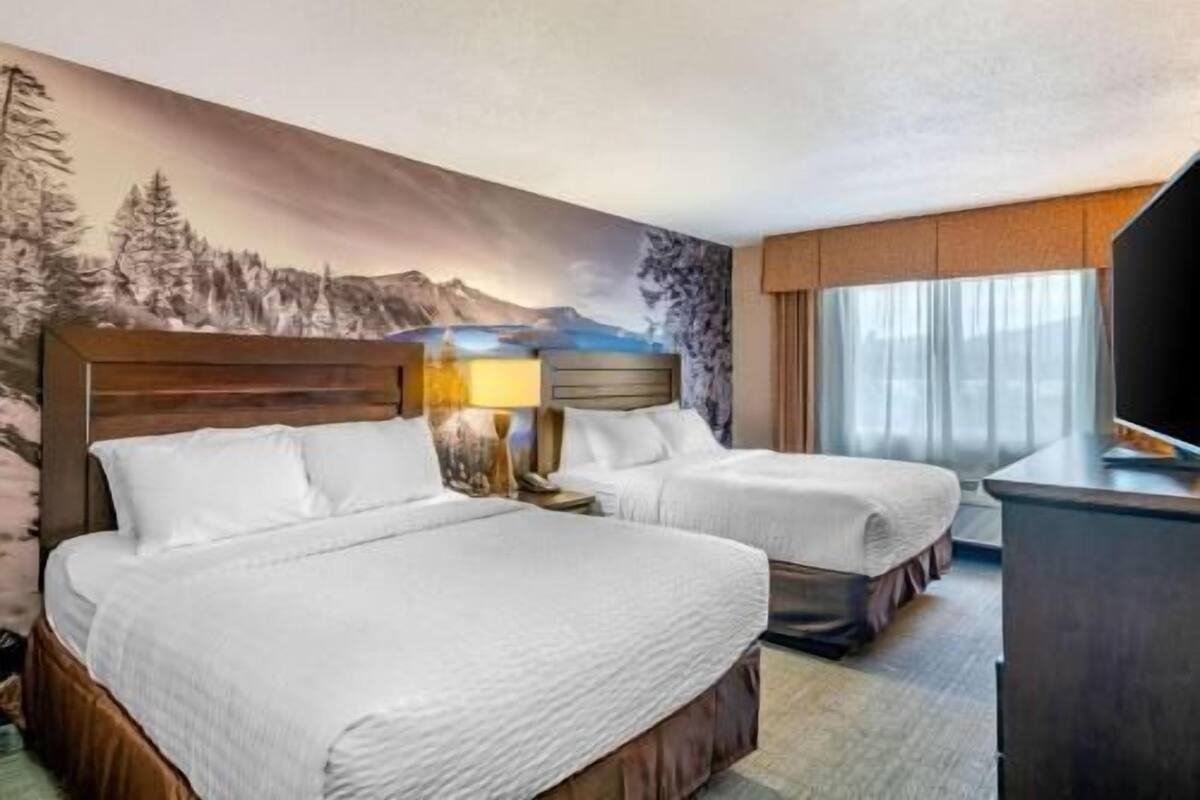 A hotel room with two beds, a nightstand with a lamp, a TV, and a mountain-themed wall mural.