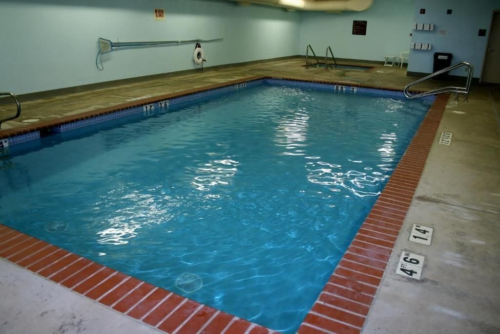 A rectangular indoor swimming pool with clear blue water, marked depth lines, and a wooden deck surrounding the pool.