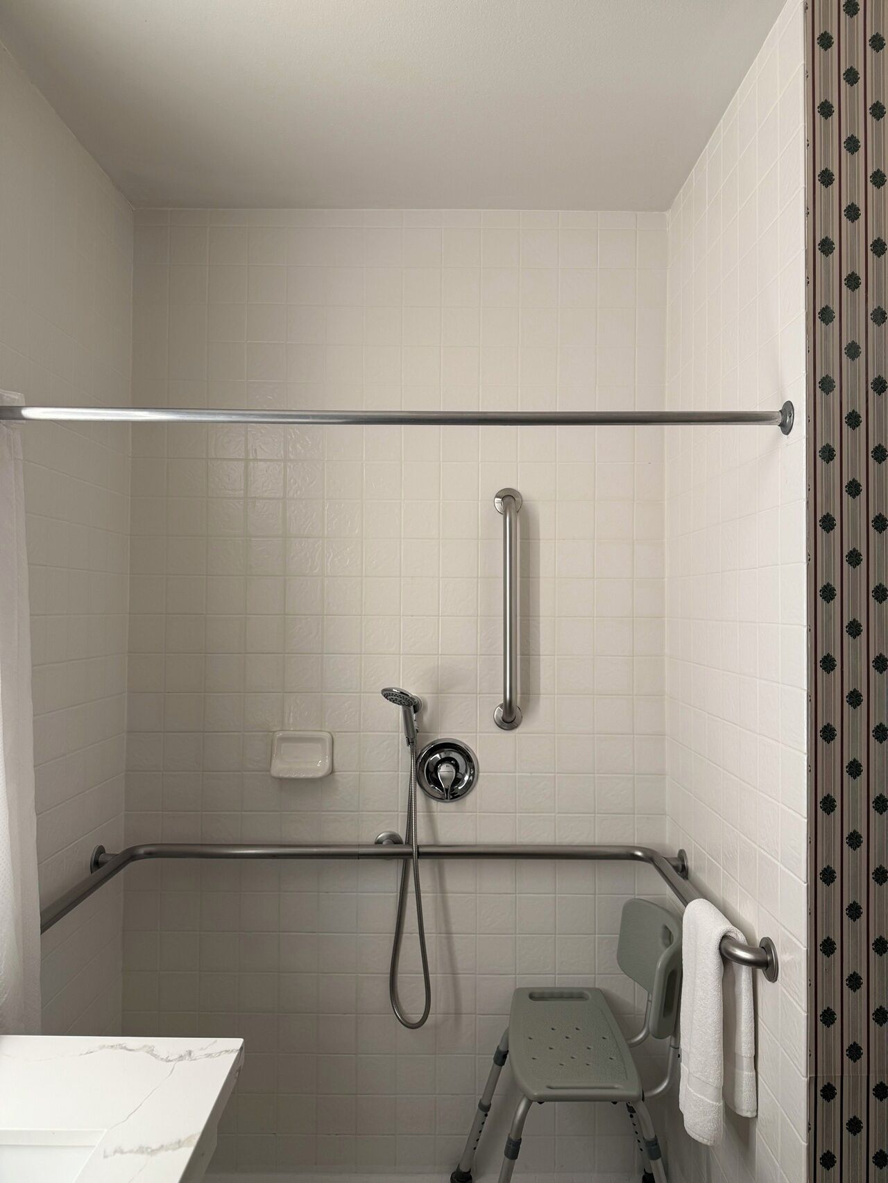 Baño con paredes de azulejos blancos, ducha con cabezal de ducha de mano, barra de apoyo, toallero y banco accesible en silla de ruedas.