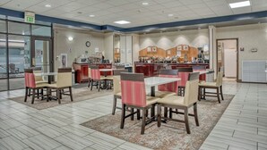 A hotel dining area with tables and chairs, a kitchen in the background, and an exit sign. - Evergreen Inn and Suites (Monroe)