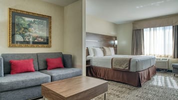 Una habitación de hotel con una cama, un sofá gris con cojines rojos, una mesa de centro de madera y un cuadro en la pared.