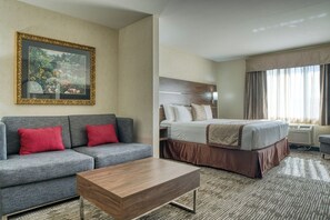 A hotel room with a bed, a gray sofa with red cushions, a wooden coffee table, and a painting on the wall.
