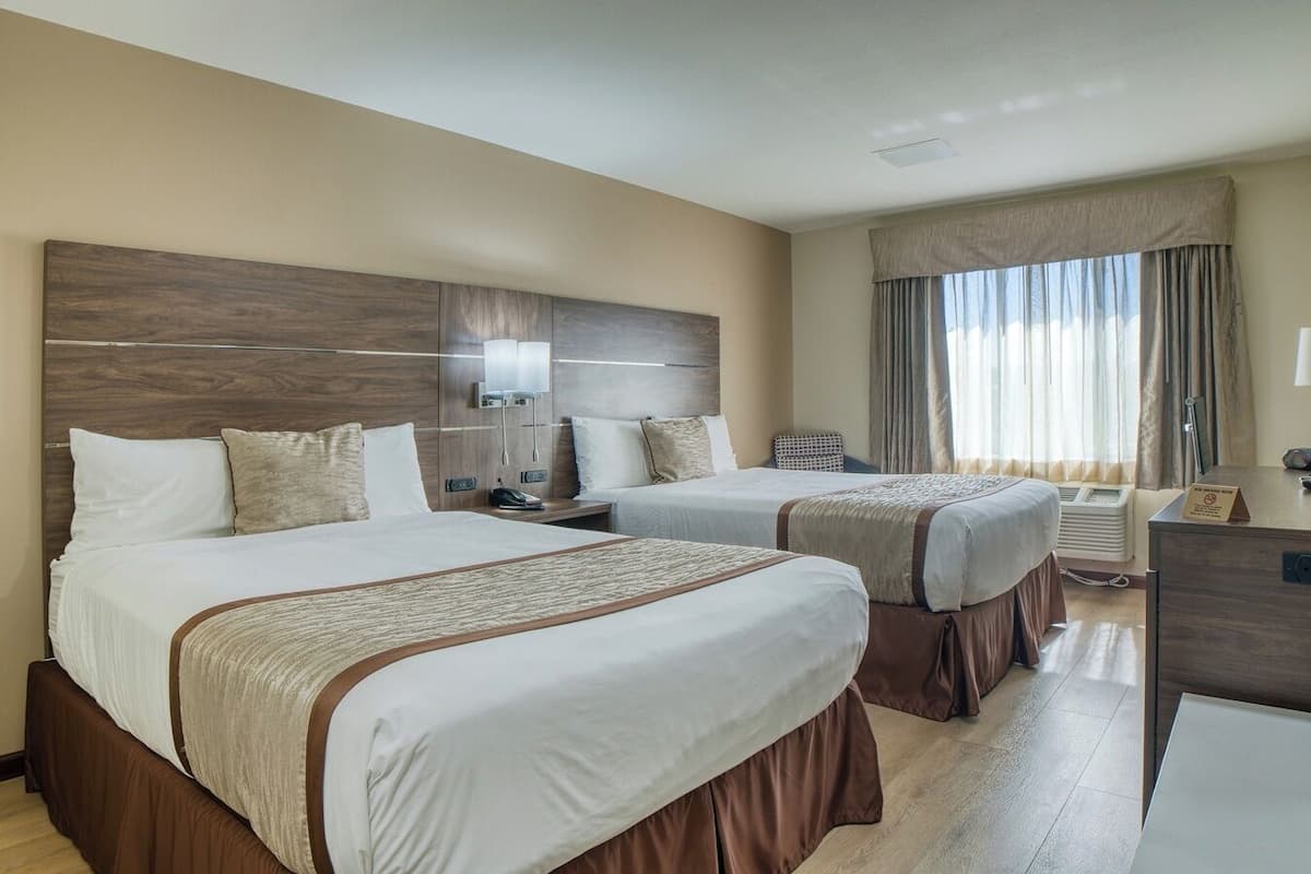 A hotel room with two beds, a desk, a lamp, and a window with curtains.