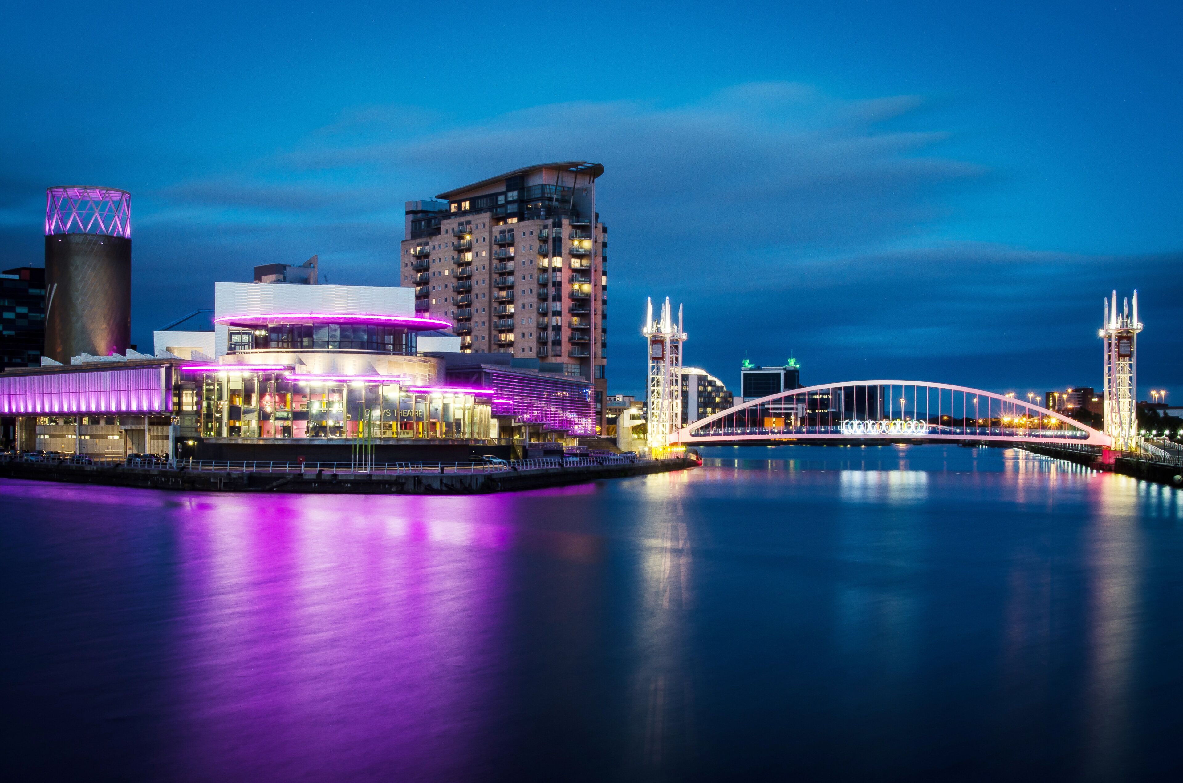 A waterfront cityscape with illuminated buildings, a bridge, and a calm river.