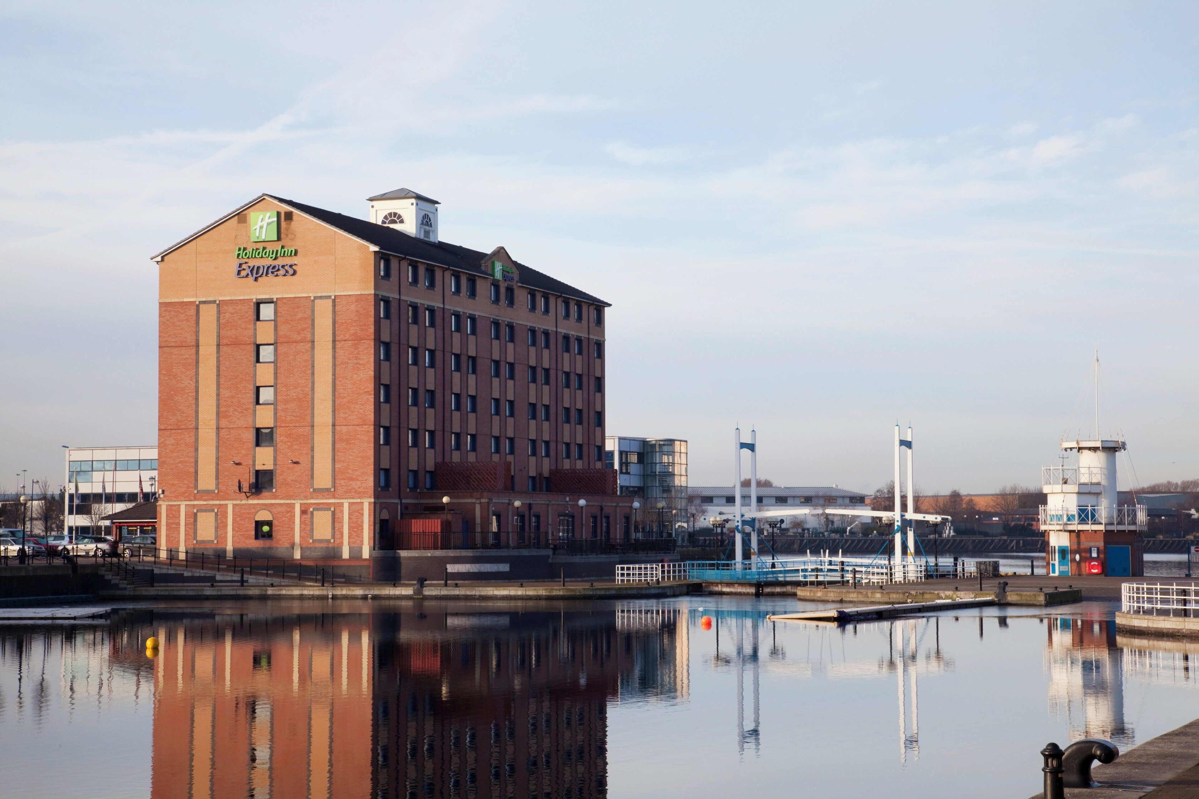 A Holiday Inn Express building with a brick facade and large windows, located near a body of water with a bridge and industrial structures in the background.