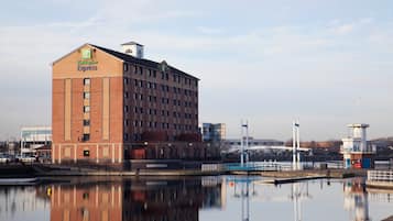 Edificio de Holiday Inn Express con fachada de ladrillo y ventanas amplias, situado cerca de una masa de agua con un puente y estructuras industriales al fondo.