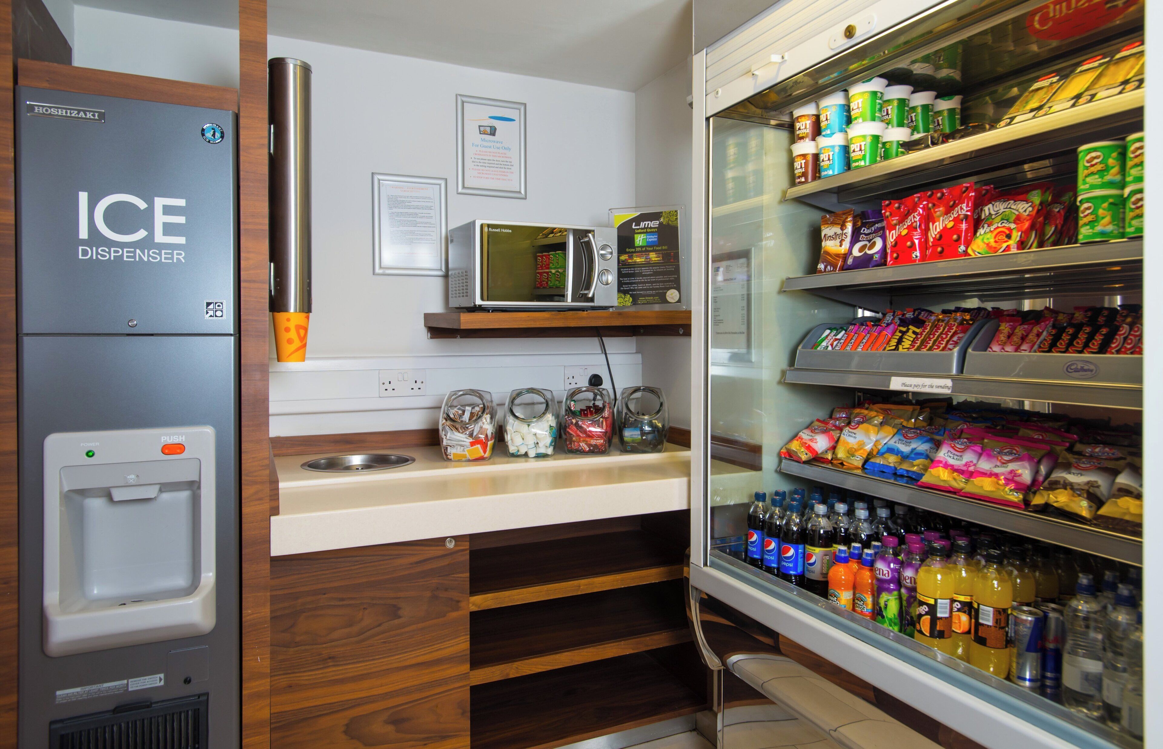 A kitchenette with an ice dispenser, microwave, and a refrigerator stocked with snacks and beverages.