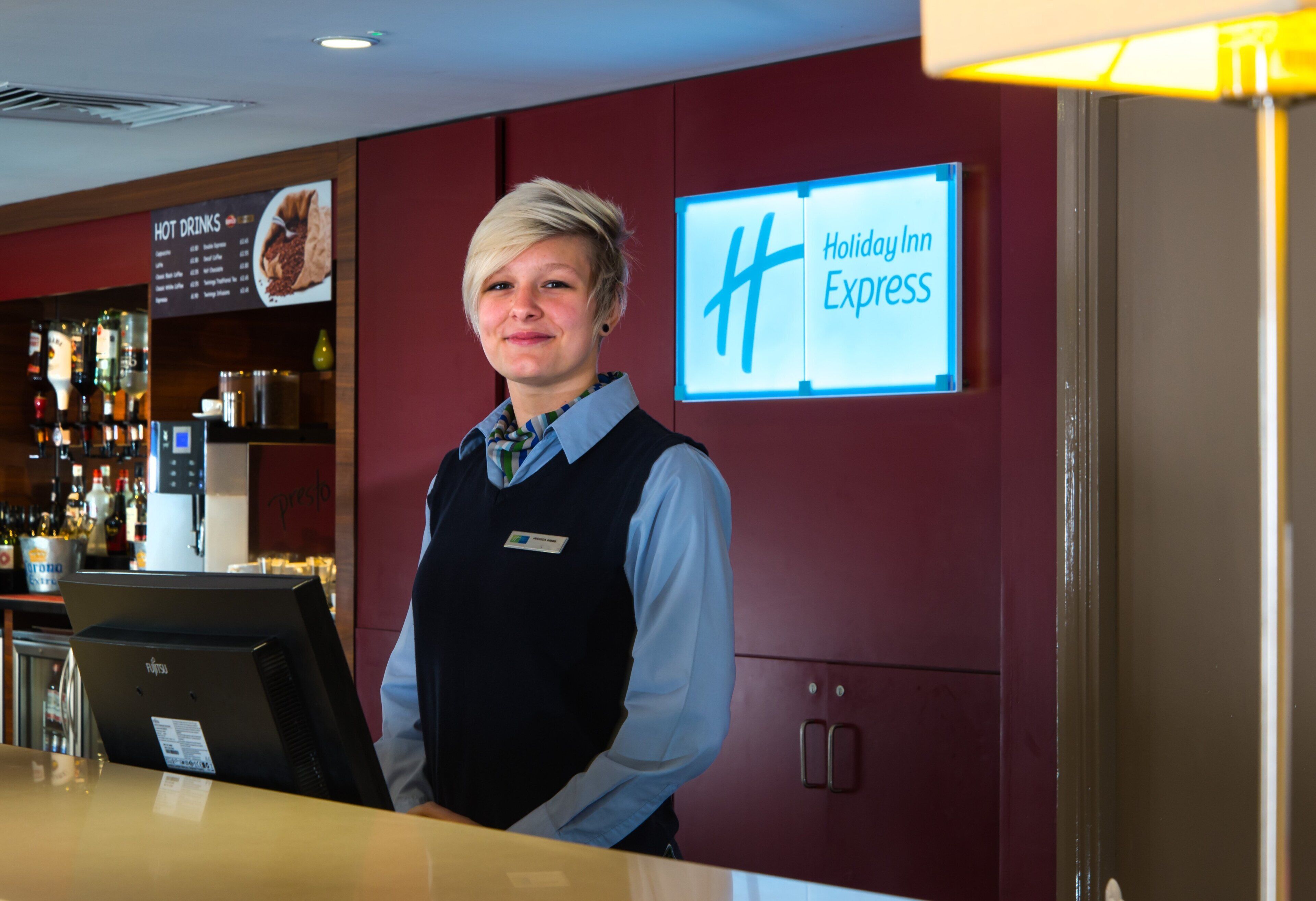 Uma área de recepção de hotel com a placa Holiday Inn Express, um bar com diversas garrafas de bebidas alcoólicas e um painel de cardápio.