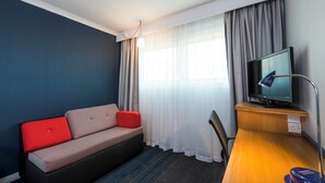 Et hotelværelse med en sofa, et skrivebord, et fjernsyn og et vindue med gardiner.